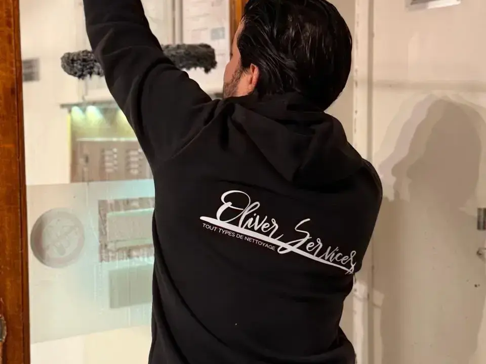 Un opérateur d'Oliver Services nettoie une vitre à la raclette, vu de dos, en veste au logo de l'entreprise.